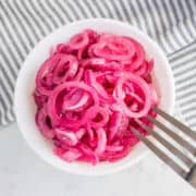 A white bowl filled with sliced pickled red onions, next to a silver fork, sits on a white surface with a striped cloth in the background.