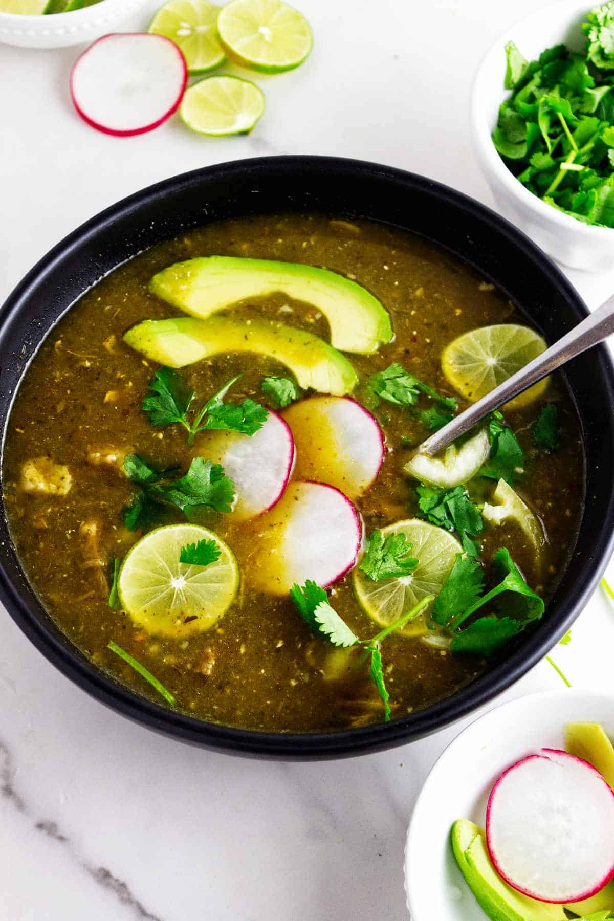 Bowl of pork pozole verde with avocado, radishes, lime, cilantro, and a spoon.
