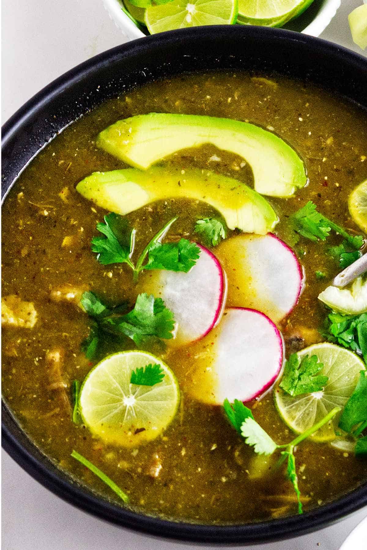 Bowl of pork pozole verde topped with avocado, radishes, lime, and cilantro.
