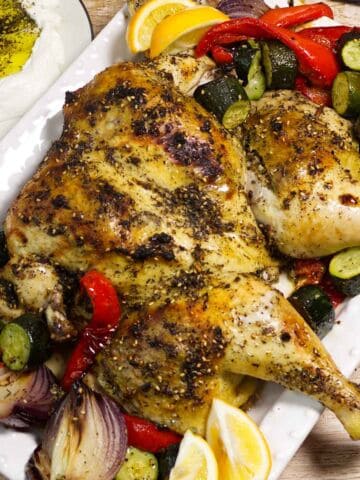 Herb-roasted spatchcock chicken with zucchini, peppers, onions, and lemon on a platter; creamy herb dip in the background.