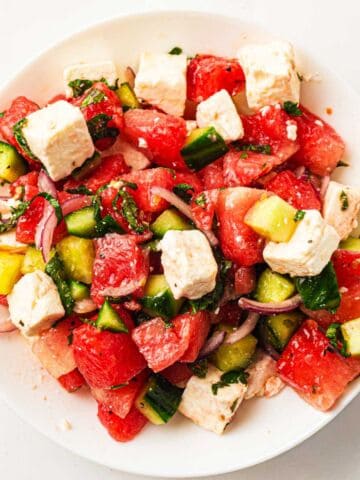 White plate with a vibrant salad of watermelon, feta, cucumber, red onion, yellow bell pepper, and herbs.