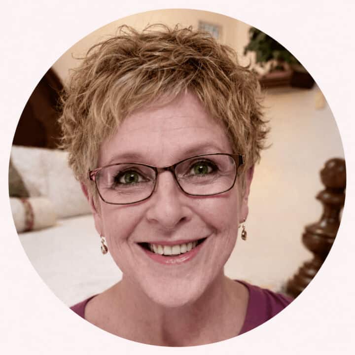 Kelly with short curly light brown hair, glasses, earrings, and a purple top sits indoors with a blurred background.