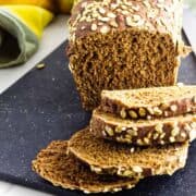 Dark brown oat bread with sliced pieces on a black board, topped with oats; yellow and green cloth in the background.