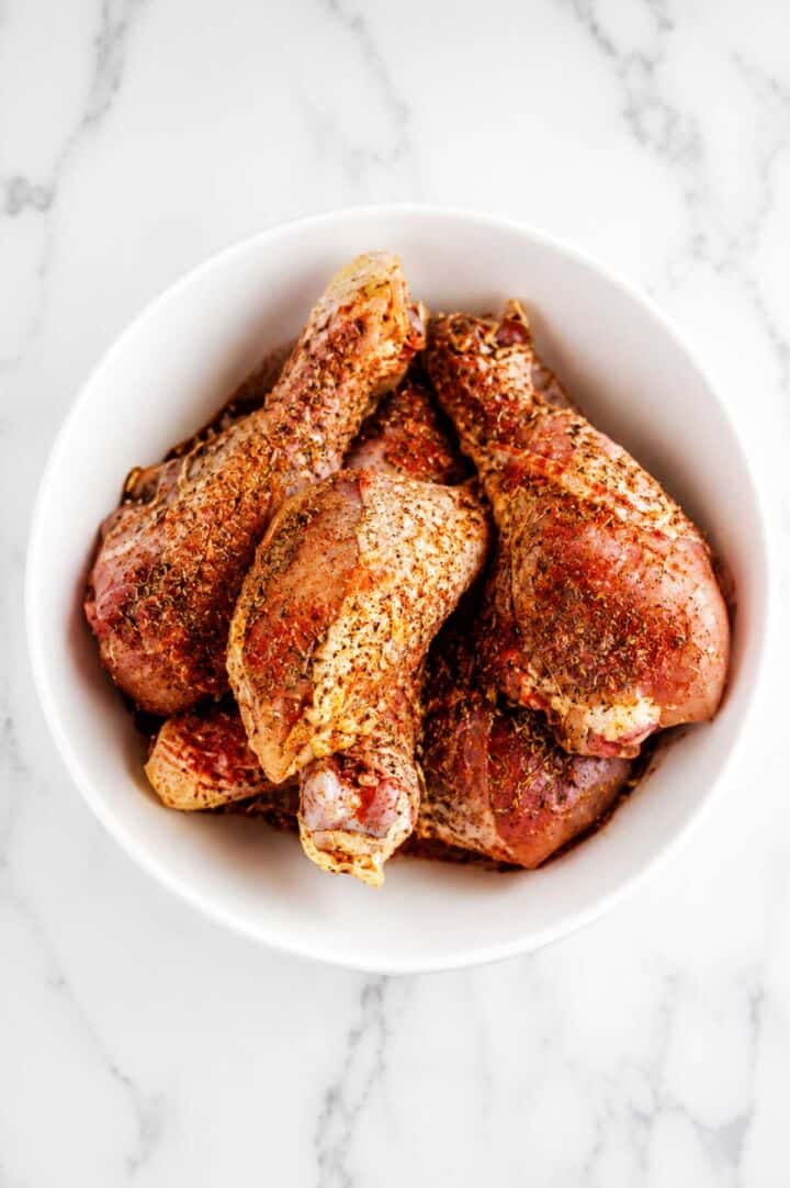 Chicken drumsticks coated with olive oil and seasonings in a white bowl before baking.