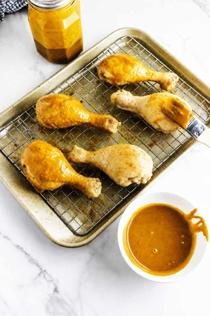 Partially baked chicken drumsticks being brushed with BBQ sauce on a wire rack over a baking sheet.