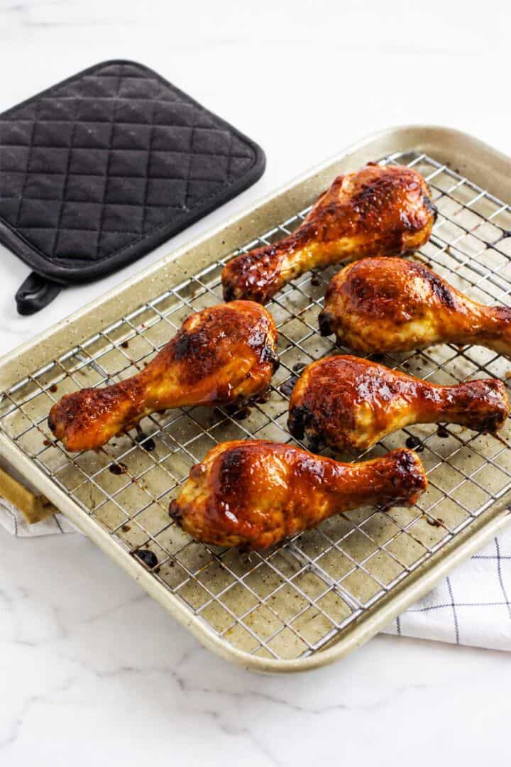 Caramelized BBQ chicken drumsticks on a wire rack over a baking sheet with a black quilted pot holder nearby.