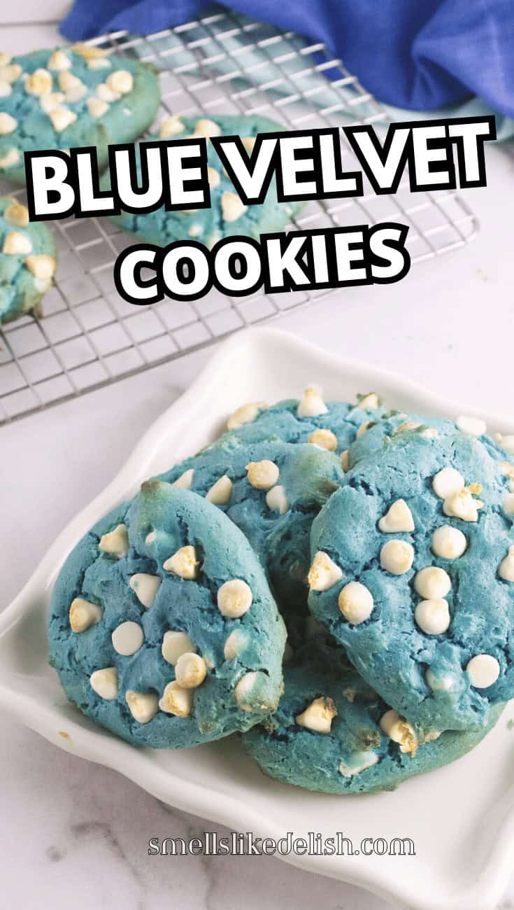 Four blue velvet cookies with white chips on a dish; more cookies, a cooling rack, and a blue cloth on a marble surface nearby.