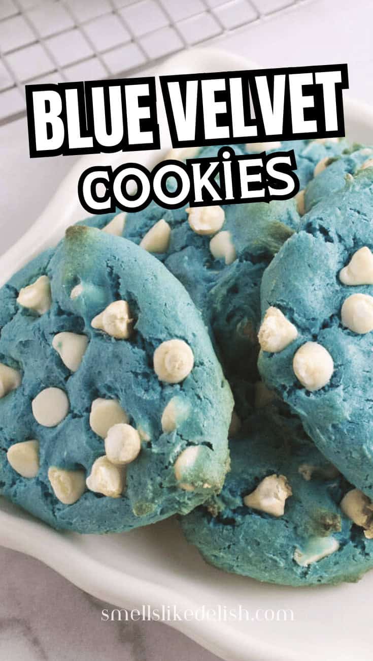 Four blue velvet cookies with white chips on a dish; more cookies, a cooling rack, and a blue cloth on a marble surface nearby.
