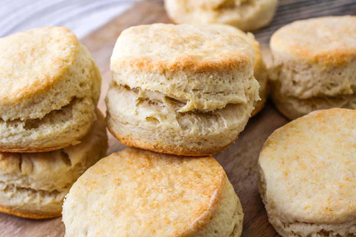 Golden brown, flaky biscuits stacked on a wooden surface, showing light, fluffy texture and crisp edges.