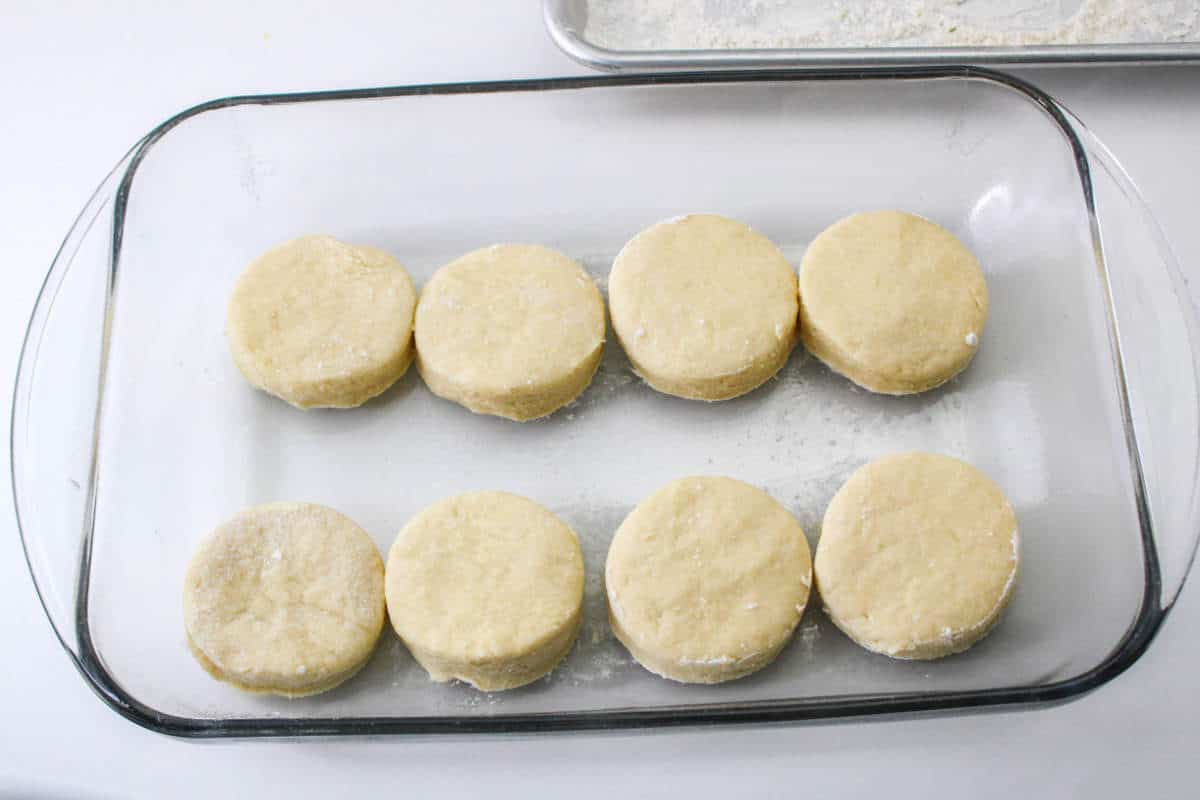 Eight unbaked biscuits in two rows sit in a glass dish on a white surface, some dusted with flour.