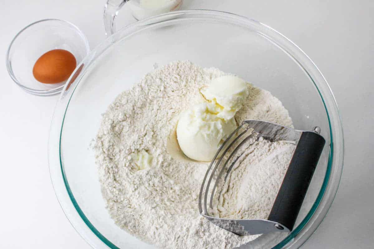 Flour and shortening are mixed in a glass bowl with a pastry blender; an egg and milk sit nearby on a white surface.
