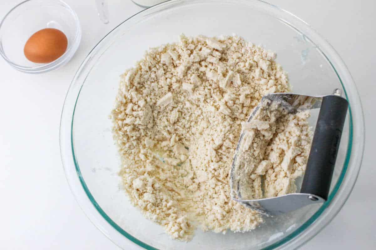 Glass bowl with crumbly flour and pastry cutter; next to it, a clear bowl holds a whole egg on a white surface.
