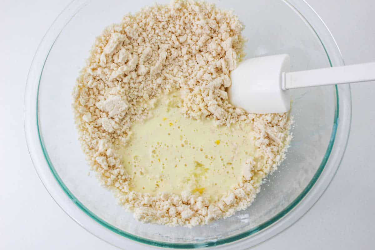 Glass bowl with dry ingredients around the edge, liquid in the center, and a white spatula inside.