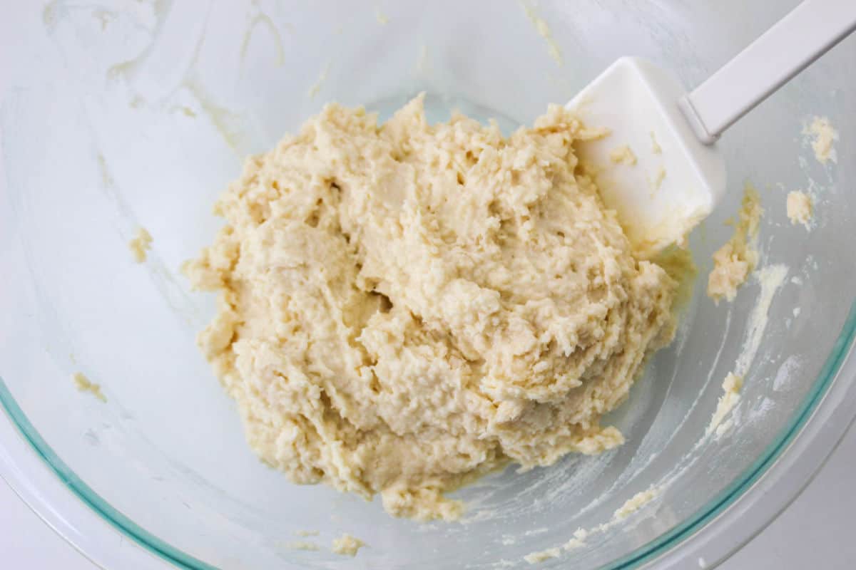 Glass bowl with pale dough and a white spatula on the side, indicating the mixture is being stirred or prepared.