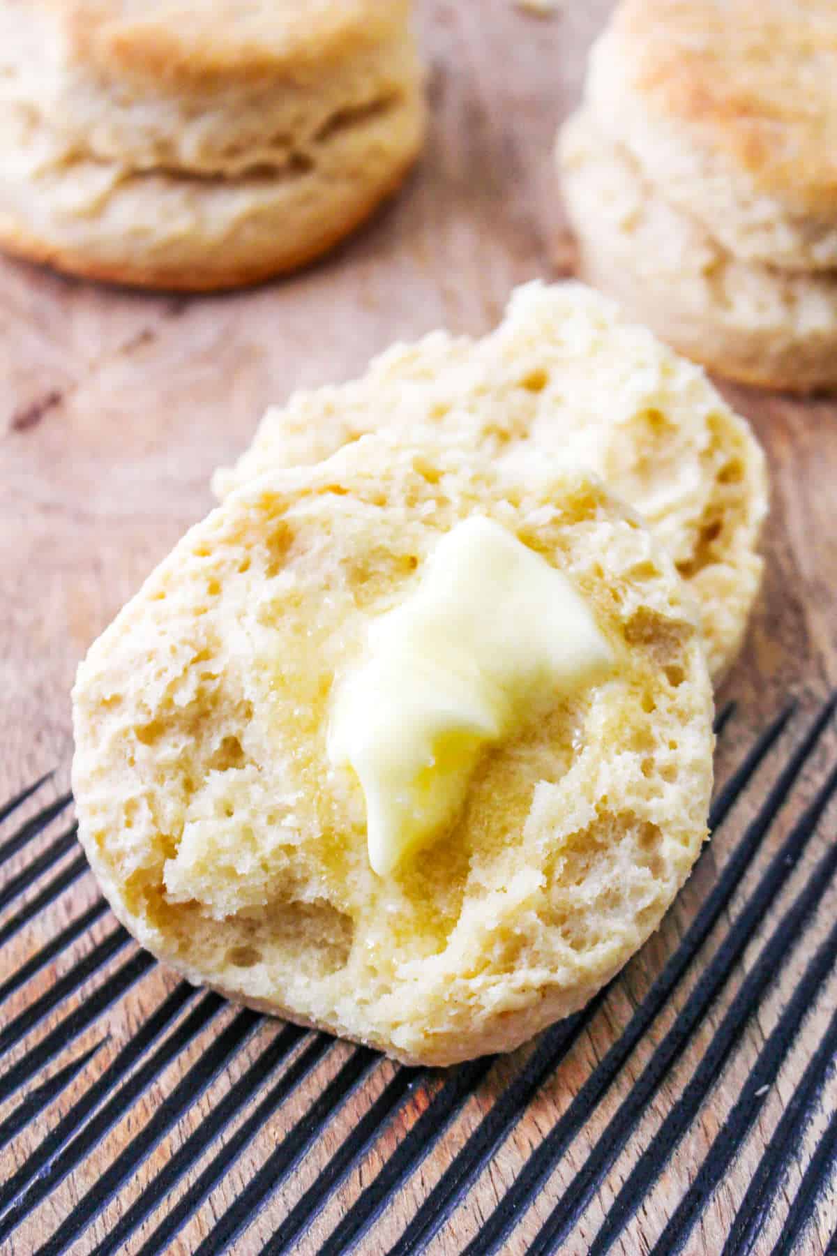 Split biscuit with melting butter in focus; two blurred biscuits behind, all on a wooden surface.