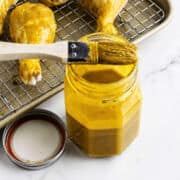 Jar of yellow BBQ sauce with basting brush beside tray of sauced raw drumsticks; jar lid on marble countertop.