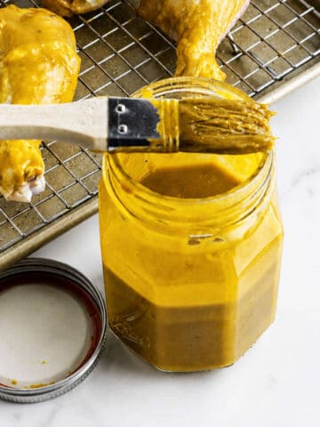 Jar of yellow BBQ sauce with basting brush beside tray of sauced raw drumsticks; jar lid on marble countertop.