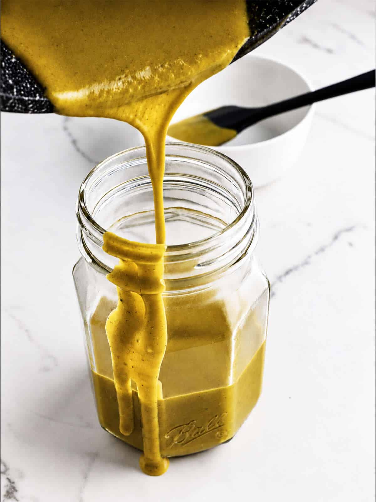 Carolina Gold BBQ sauce pouring from a saucepan into a glass jar on white marble.