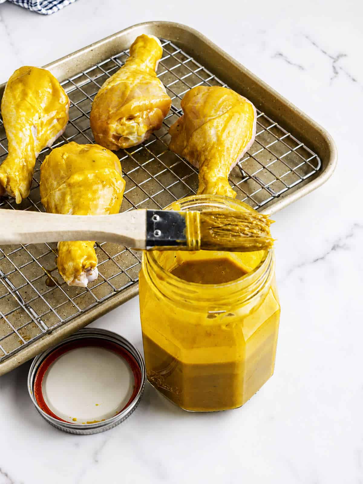 Brush resting on a jar of Carolina Gold BBQ sauce beside sauced chicken drumsticks on a wire rack.