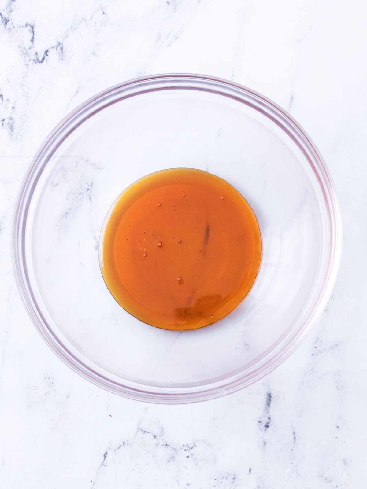 Glass mixing bowl with a small amount of amber liquid on a white marble surface.