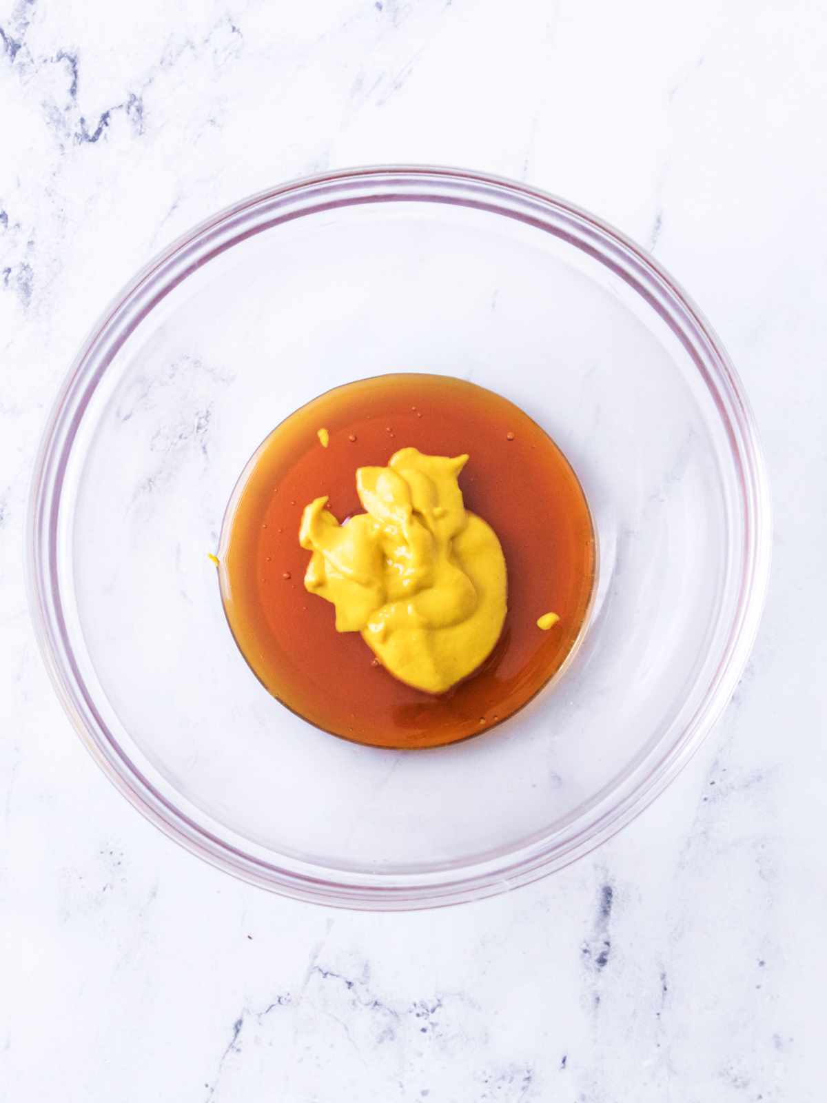 Glass bowl with yellow mustard and brown honey on a white marble surface.