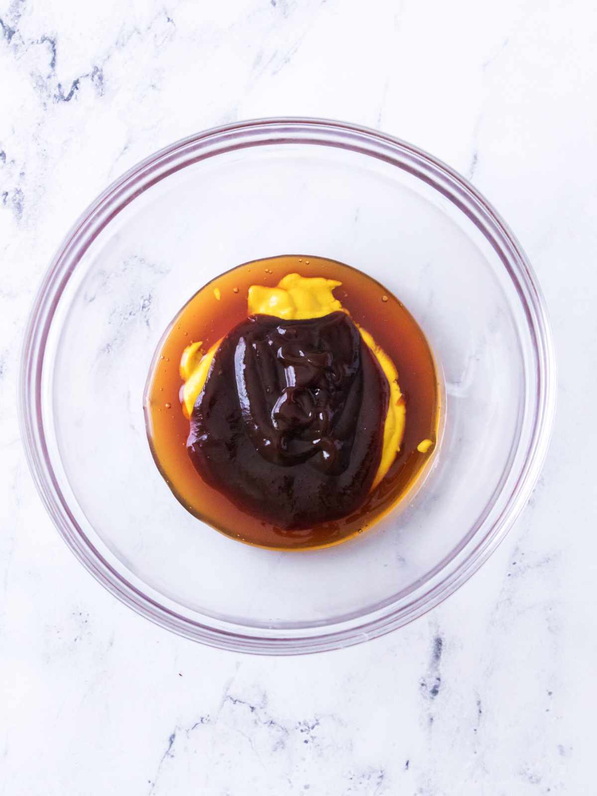 Glass bowl on white marble holds yellow mustard, brown barbecue sauce, and dark soy sauce.