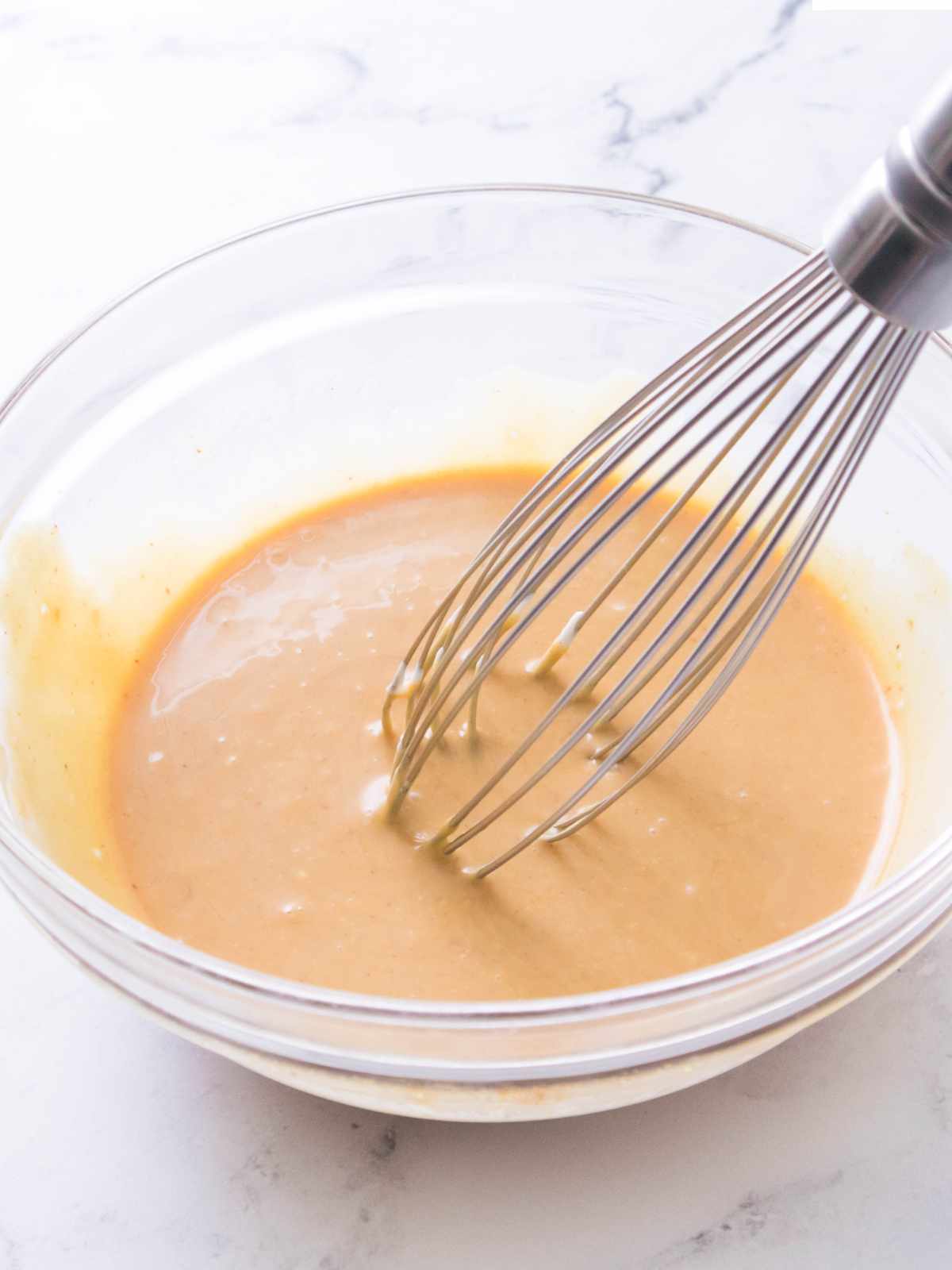 A metal whisk stirs light brown batter in a clear glass bowl on a white marble countertop.