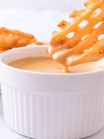 Waffle fry dipped in creamy sauce in a white ramekin, with other waffle fries blurred in the background.