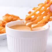 Hand dipping a waffle fry into a white ramekin of creamy sauce, with extra fries on a plate in the background.