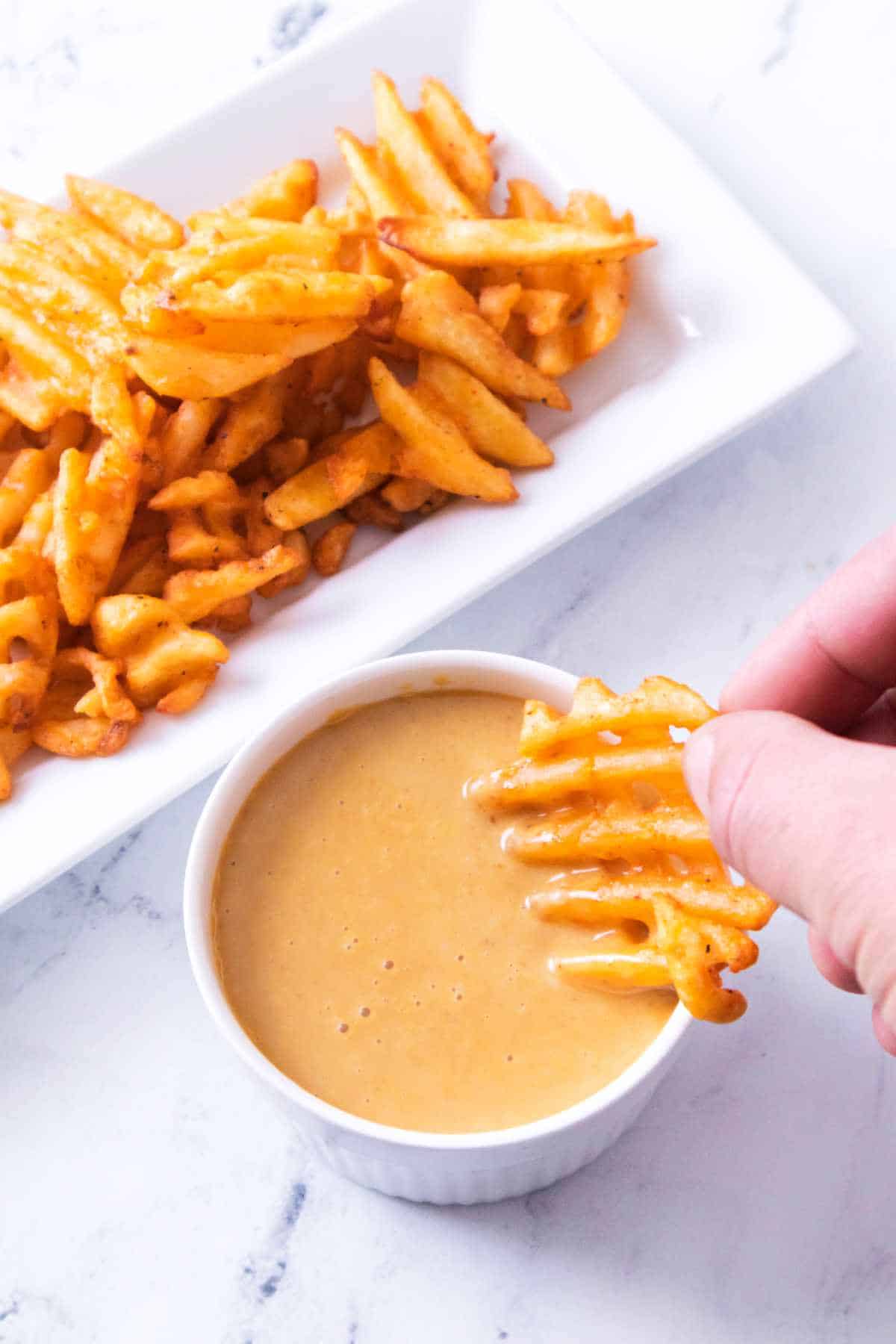A hand dips a waffle fry into creamy sauce, with extra fries on a rectangular plate seen in the background.