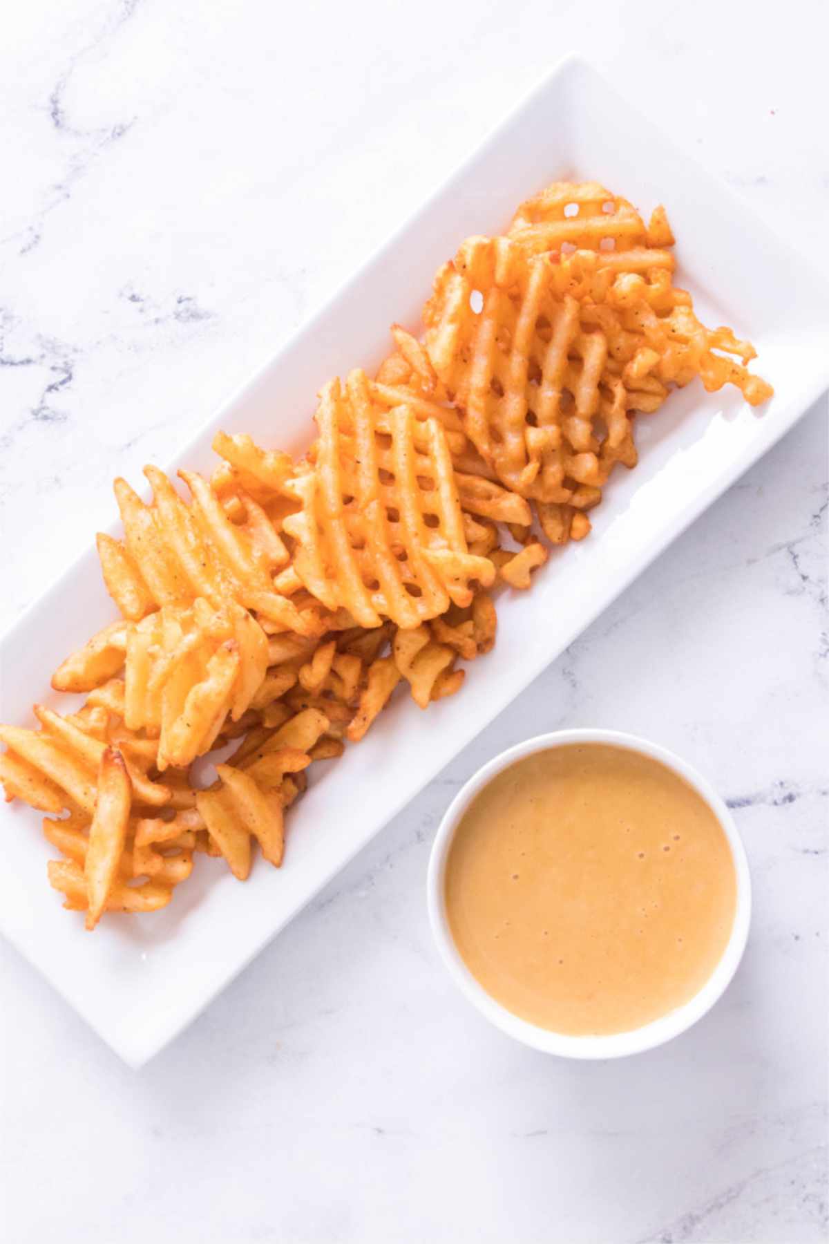 Waffle fries on a white plate with a small bowl of light brown dipping sauce, set on a white marble surface.