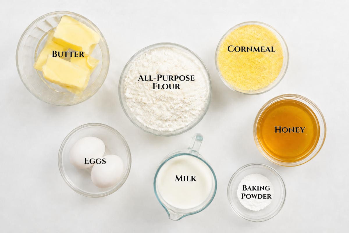 Seven bowls with labeled baking ingredients: butter, flour, cornmeal, honey, eggs, milk, and baking powder.