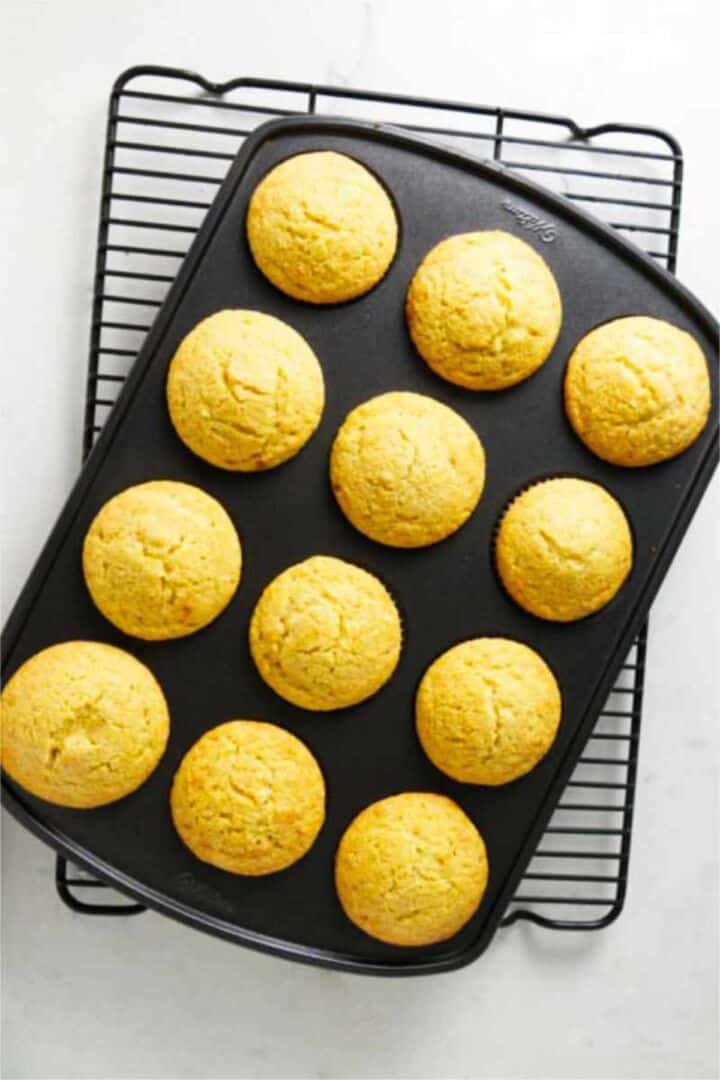 Twelve golden cornbread muffins cooling in a muffin tin on a wire rack.