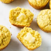Golden cornbread muffins on a white surface, one split open with melting butter. The tops are rough and the texture is crumbly.
