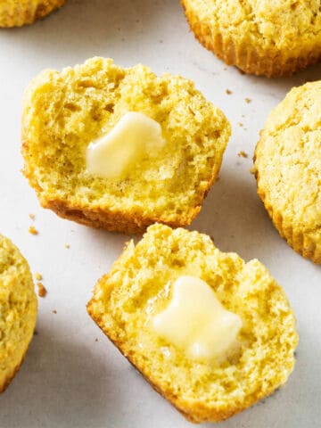 Golden cornbread muffins on a white surface, one split open with melting butter. The tops are rough and the texture is crumbly.