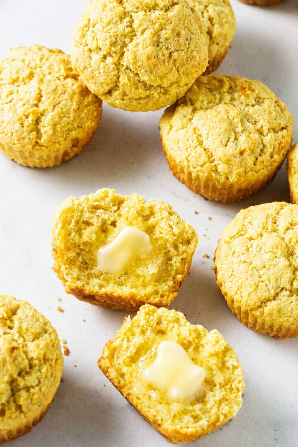 Golden cornbread muffins on a white surface with one split open and butter melting inside.