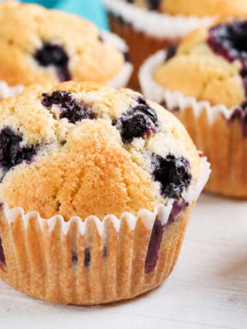 Fresh dairy-free blueberry muffins with golden tops in white liners, juicy berries showing, on a light-colored surface.