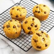 Five golden brown gluten-free chocolate chip muffins cool on a black wire rack atop white marble, chips visible on top.