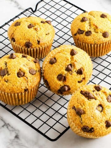 Five golden brown gluten-free chocolate chip muffins cool on a black wire rack atop white marble, chips visible on top.