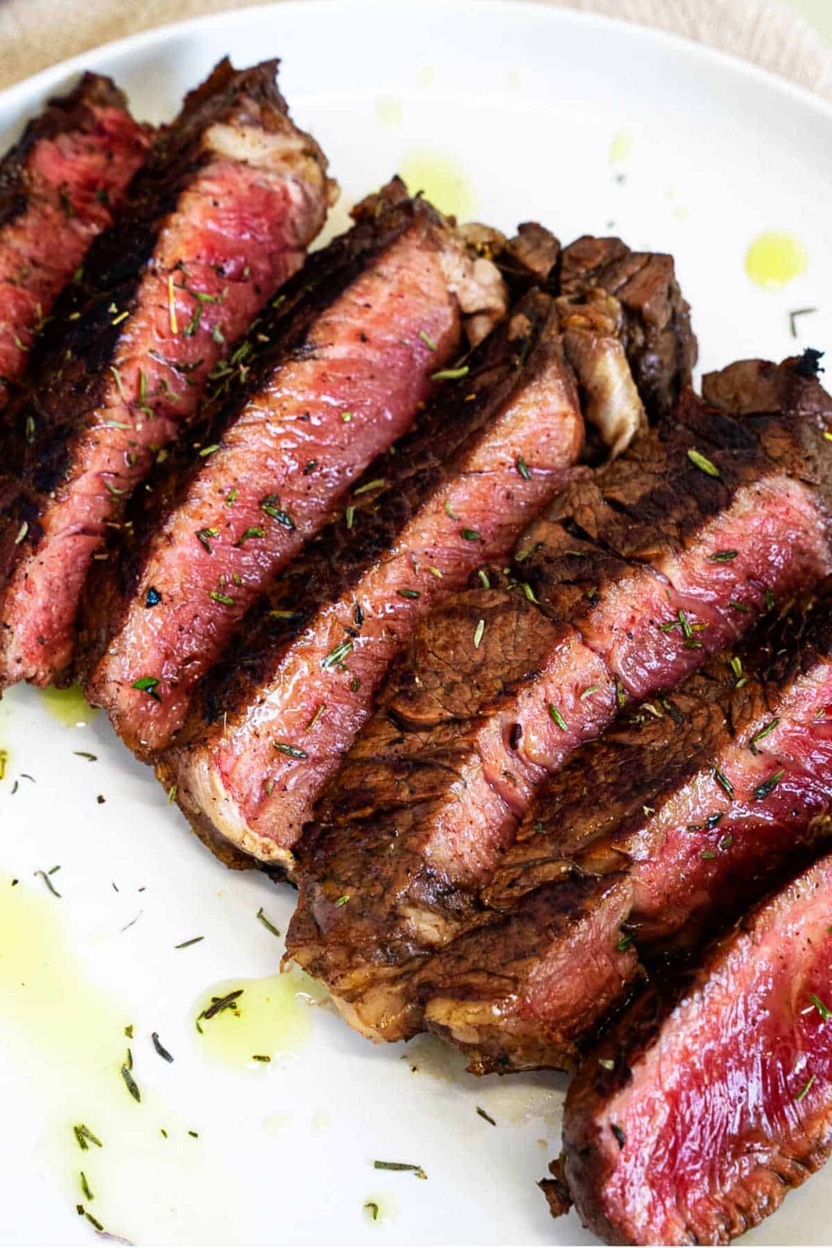 Sliced grilled top sirloin steak on a white plate with herbs, olive oil, seared edges, and a pink center.