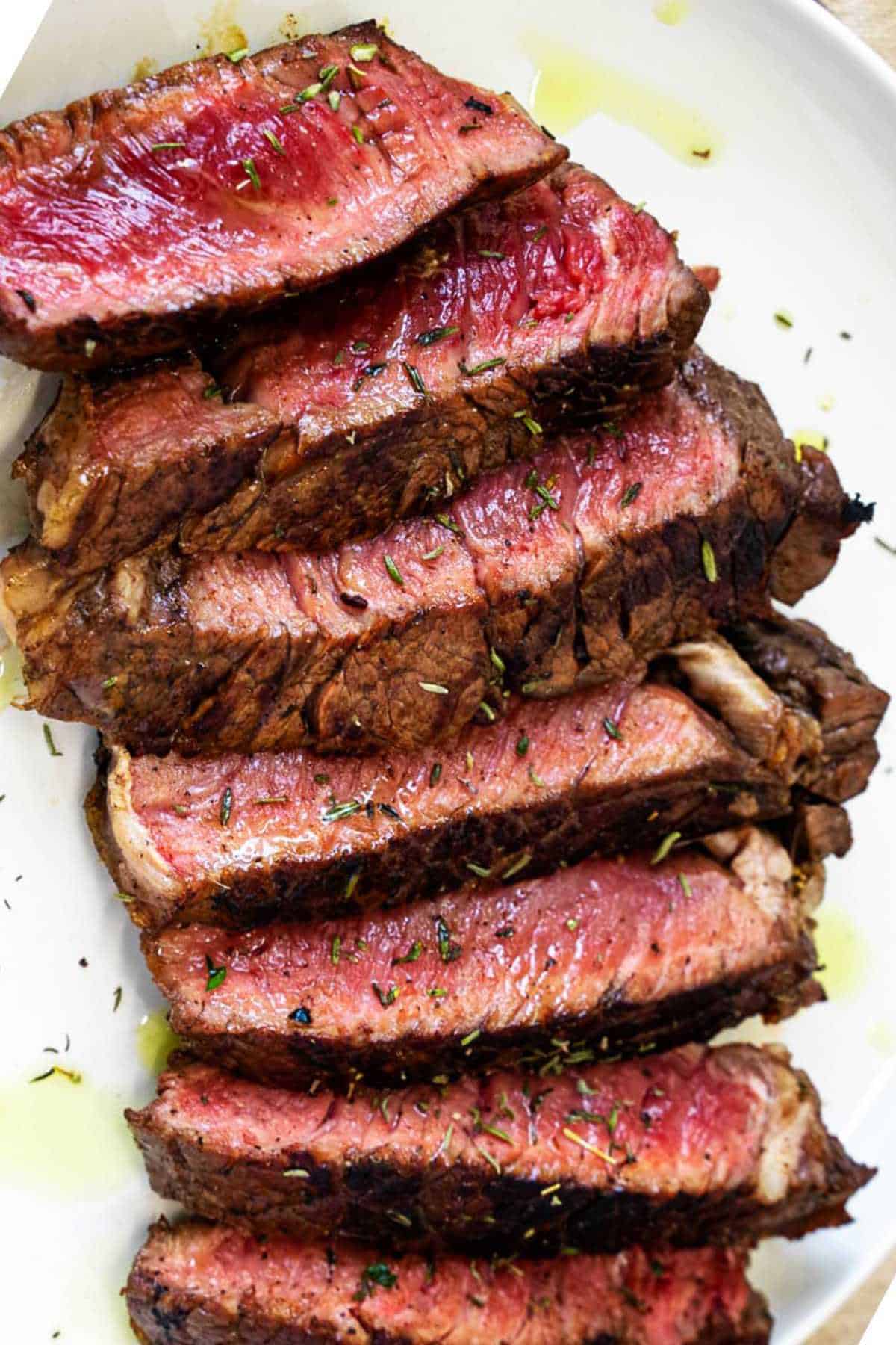 Sliced grilled top sirloin steak on a white plate with herbs, olive oil, and a juicy pink center.
