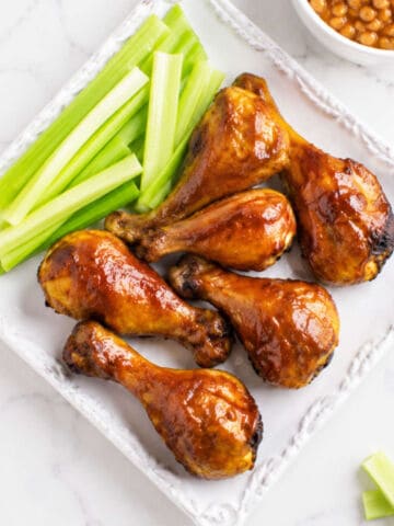 Six BBQ chicken drumsticks and celery sticks on a white plate; baked beans partially visible in the corner.