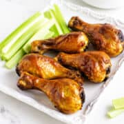 Six glazed BBQ chicken drumsticks and fresh celery sticks on a white plate against a plain white background.