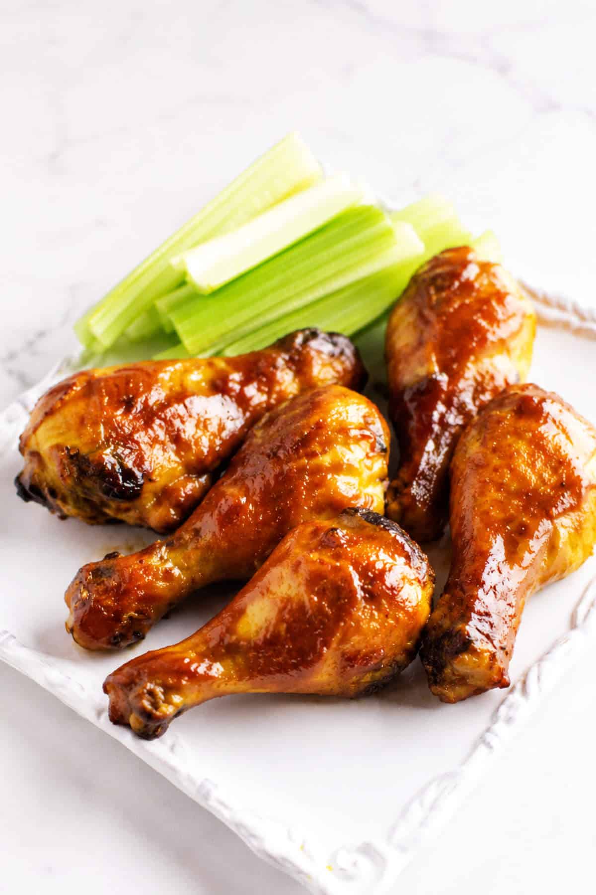 Oven baked BBQ chicken drumsticks and celery sticks on a white plate on a marble surface.