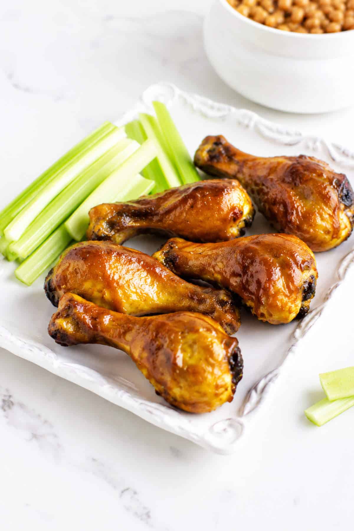 BBQ chicken drumsticks and celery sticks on a white plate with baked beans in the background.