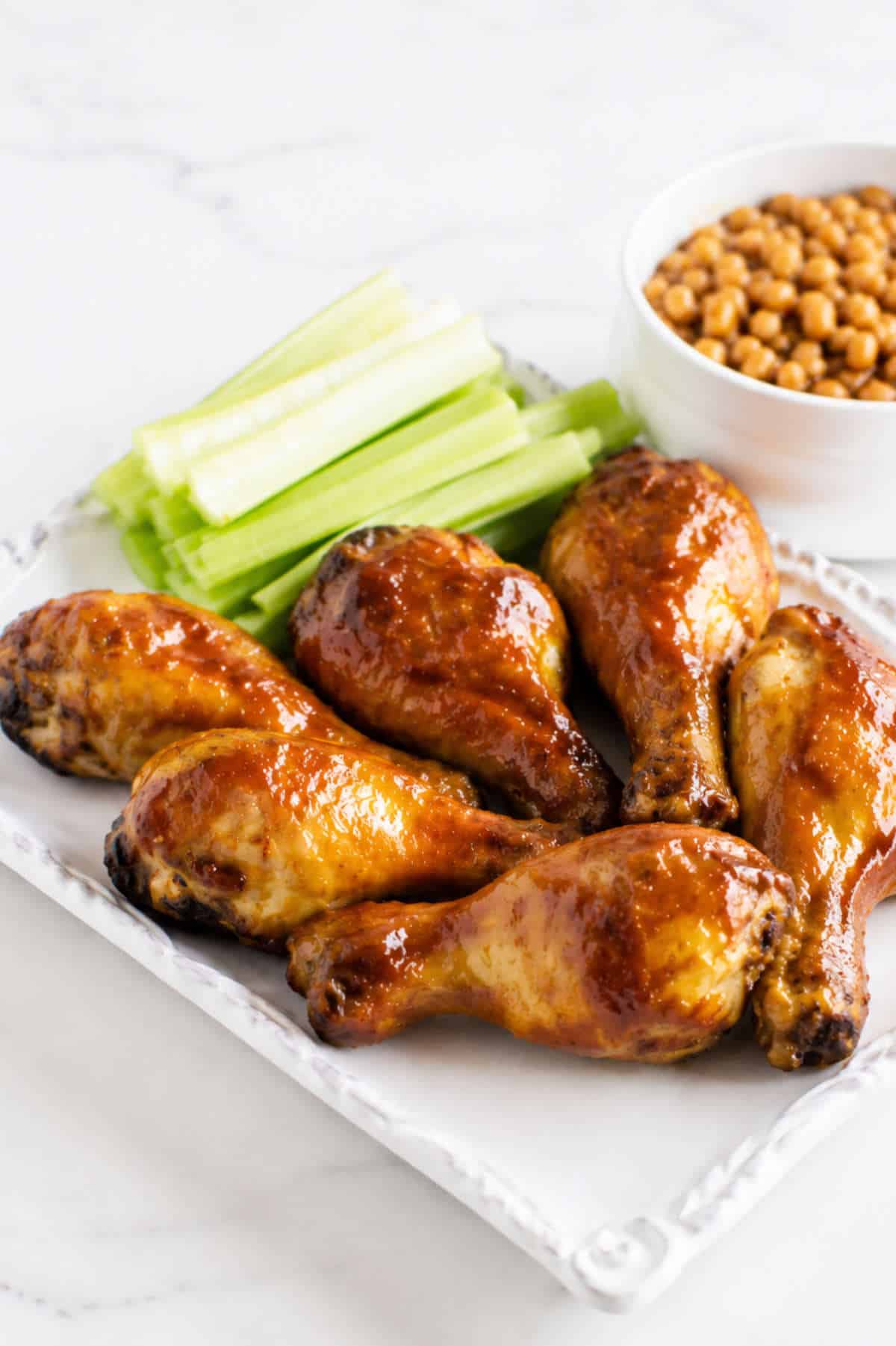 BBQ chicken drumsticks with celery sticks on a plate and baked beans served on the side.