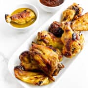 White plate of golden-brown Honey Gold chicken wings with mustard and barbecue sauces, set on a white surface.