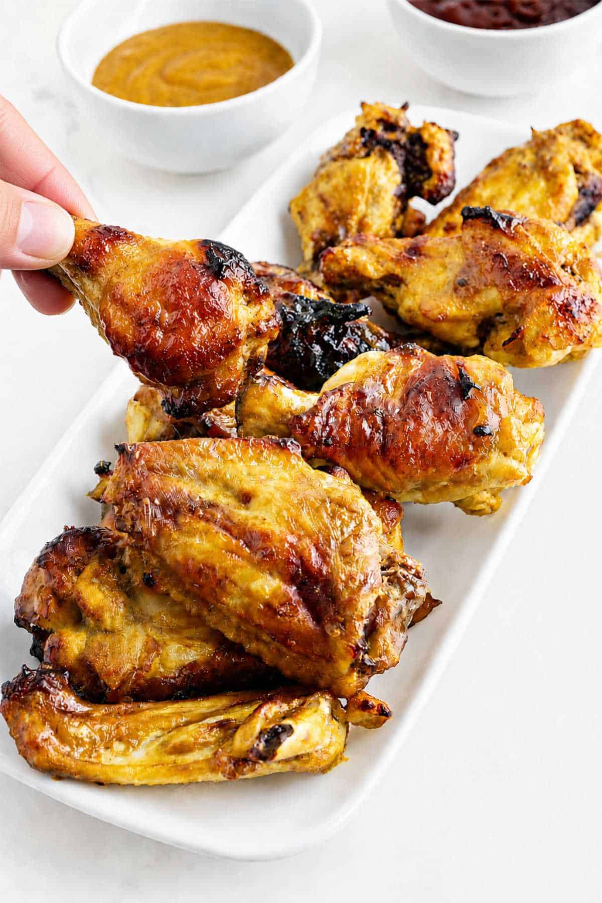 Hand holding a Honey Gold wing over a platter of golden BBQ chicken wings with dipping sauces in the background.