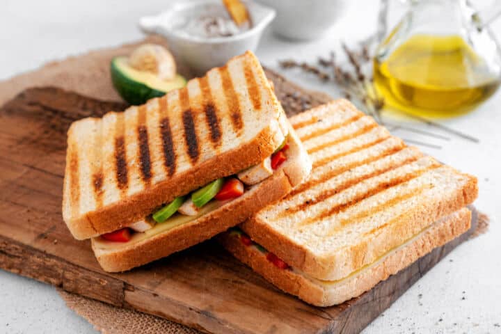 Grilled Panera Chipotle Chicken Avocado Melt sandwiches on a wooden board with light grill marks and sweet red peppers showing.