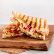 Grilled sandwich halves with cheese, tomato, and avocado on a wooden board; bread has clear grill marks.